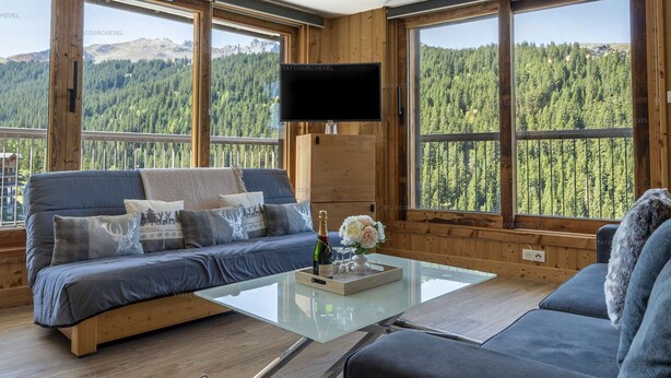 A modern living room with a sofa, a glass coffee table, and a TV mounted on the wall. Large windows offer a view of a forested mountain area. - Ski-in/ski-out apartment for families with balcony and parking (Courchevel 1650)