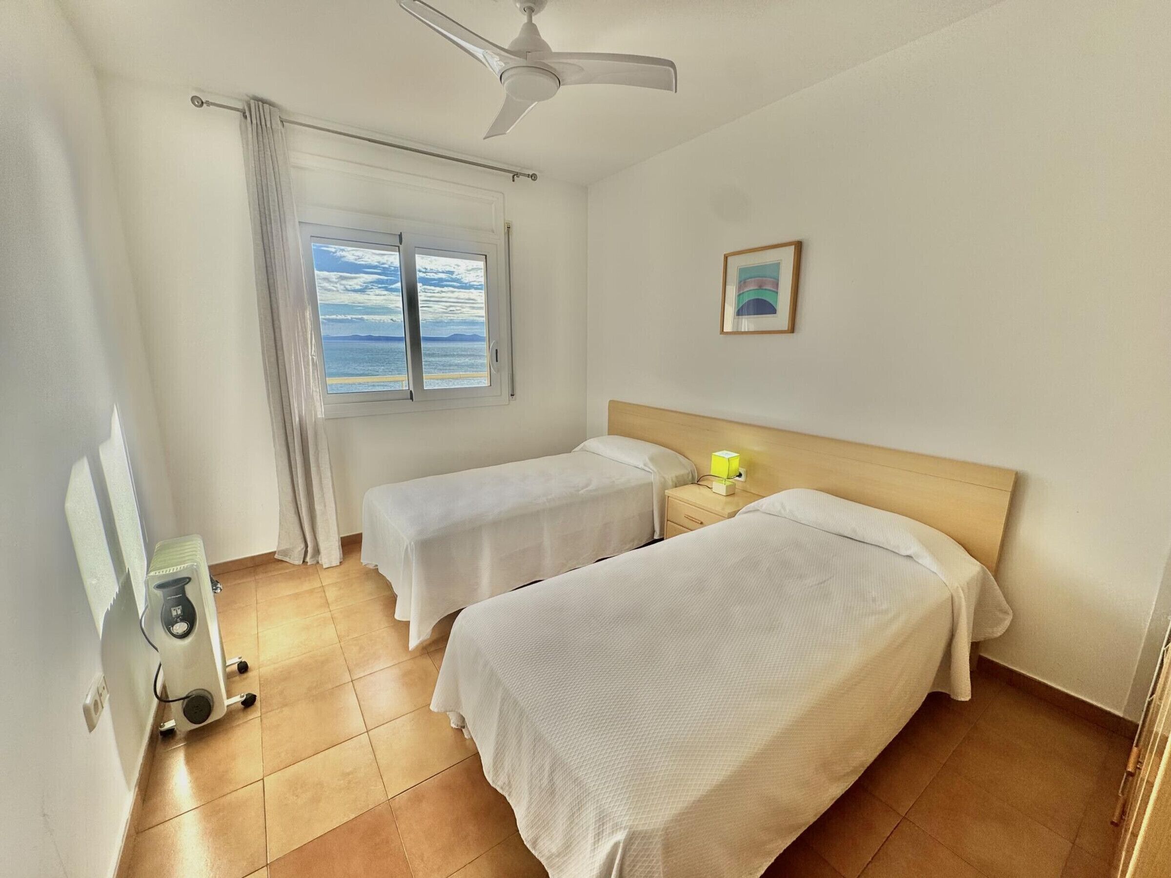 Hotellihuone, jossa on kaksi sänkyä, kattotuuletin, ikkuna, josta on näkymä merelle, sekä yöpöytä, jossa on lamppu.
