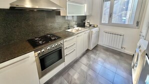 A modern kitchen with a gas stove, oven, white cabinets, and a window with a view of buildings. - Spacious Apartment in Central Roses, 650m from Beach, with Terrace & Private Parking (Roses)