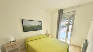 A bedroom with a bed, a nightstand, a lamp, a painting, and a view through a window. - Spacious Apartment in Central Roses, 650m from Beach, with Terrace & Private Parking (Roses)