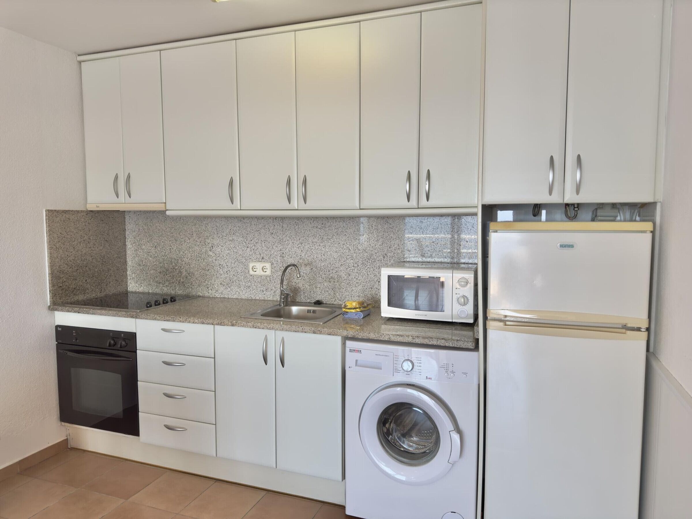 Uma cozinha compacta com armários brancos, micro-ondas, máquina de lavar e lava-louças.
