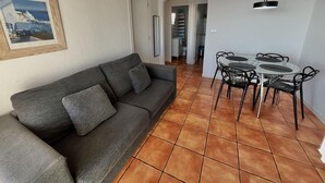 Un salon avec un canapé gris foncé, une table à manger en verre entourée de chaises et un sol carrelé.