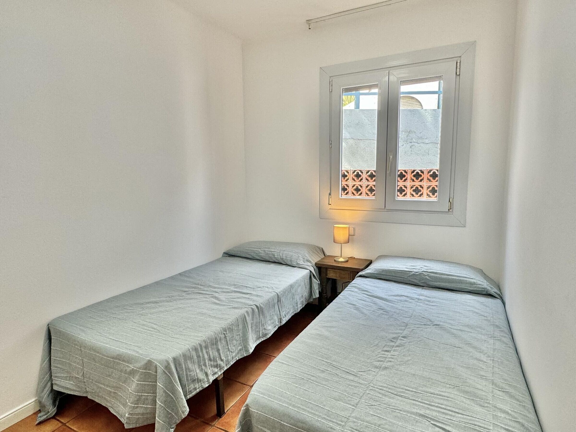 A small, clean room with two single beds, a window with decorative tiles, and a bedside lamp.