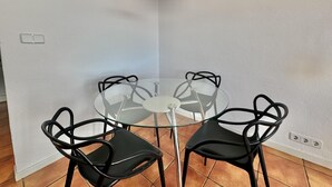 Un coin repas avec une table en verre et quatre chaises noires, un luminaire suspendu noir et un mur blanc doté d’une prise électrique.