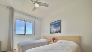 Una habitación de hotel con dos camas, un ventilador de techo, una ventana con cortinas, una mesita de noche con una lámpara y un cuadro en la pared.