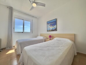 Ein Hotelzimmer mit zwei Betten, einem Deckenventilator, einem Fenster mit Vorhängen, einem Nachttisch mit Lampe und einem Gemälde an der Wand.