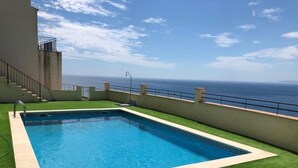 Una piscina rectangular con superficie de agua azul cristalina, rodeada de césped artificial y vista al océano.