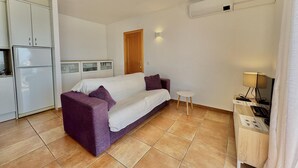 A compact room with a sofa, a small table, a TV, and a kitchen area including a fridge and cabinets. - Apartment with pool, AC, and parking 700m from the beach in Roses (Roses)
