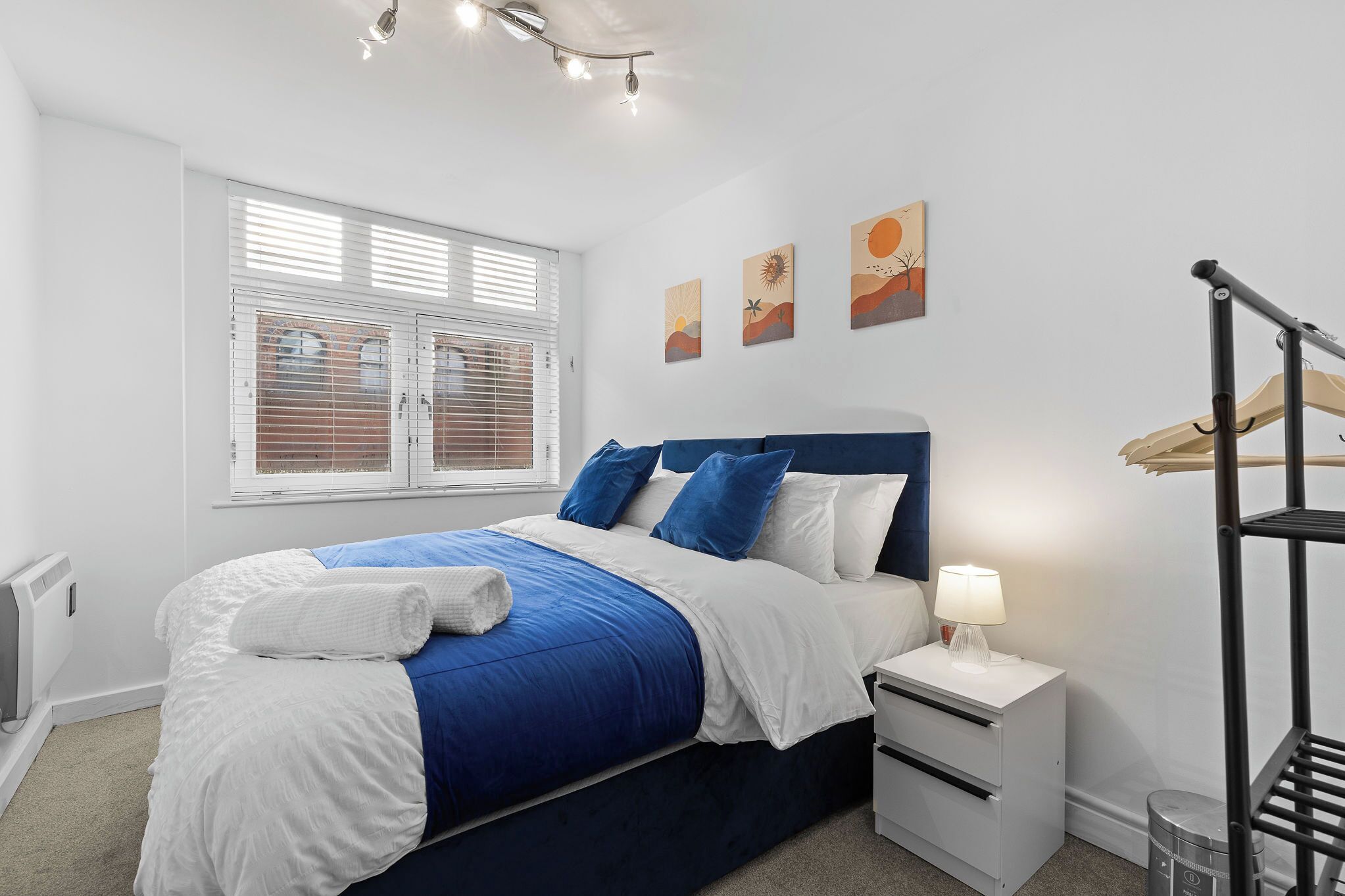 Un dormitorio ordenado con cama, mesita de noche, lámpara y ventana con persianas.