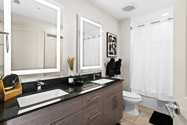 Een moderne badkamer met een dubbele wastafel, een toilet, een douche met een witte douchegordijn en een grote spiegel boven de wastafel.