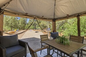 A covered outdoor seating area with a table and chairs, a hammock, and a view of the surrounding forest. - Lovely 2-bedroom cabin bordering Zion's national Park, Close to cable Mountain. (Orderville)