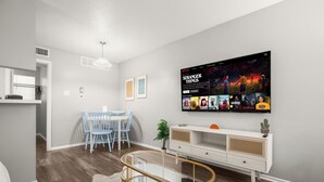 A modern living room with a flat-screen TV displaying 'Stranger Things' on Netflix, a dining area with blue chairs, a glass coffee table, and a white console table with decorative items. - Modern Central Austin Retreat Near UT & Downtown (Austin)
