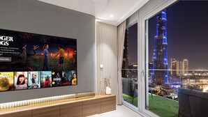 Un salon moderne doté d’un grand téléviseur diffusant des affiches de films, d’une console en bois, d’un vase blanc et offrant une vue sur le paysage urbain illuminé la nuit.