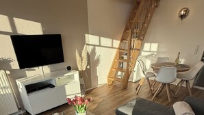 A modern living room with a large flat-screen TV, a wooden coffee table, a grey sectional sofa, and a dining area with a wooden staircase. - Dat Stürboord with North Sea view, swimming pool and sauna (Wurster Nordseeküste)