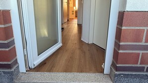 A hallway with a wooden floor leading to a room with a door. There is a doormat with a house symbol and the text "Hereinspaart". - Dat Stürboord with North Sea view, swimming pool and sauna (Wurster Nordseeküste)