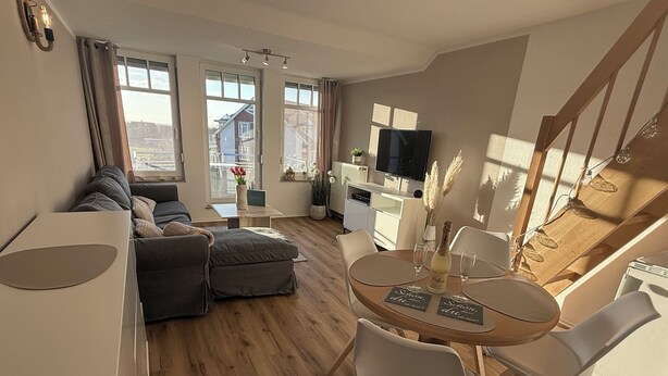 A modern living room with a sofa, ottoman, dining table with chairs, a flat-screen TV, and a kitchen area with a refrigerator and microwave. - Dat Stürboord with North Sea view, swimming pool and sauna (Wurster Nordseeküste)