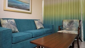 A living room with a blue sofa, a wooden coffee table, and a chair with a leaf-patterned cushion. A framed painting is on the wall. - Last minute deal on Aruba Resort;Large 1st Floor Suite;Sleeps 4; 1 MasterBedroom (Oranjestad)