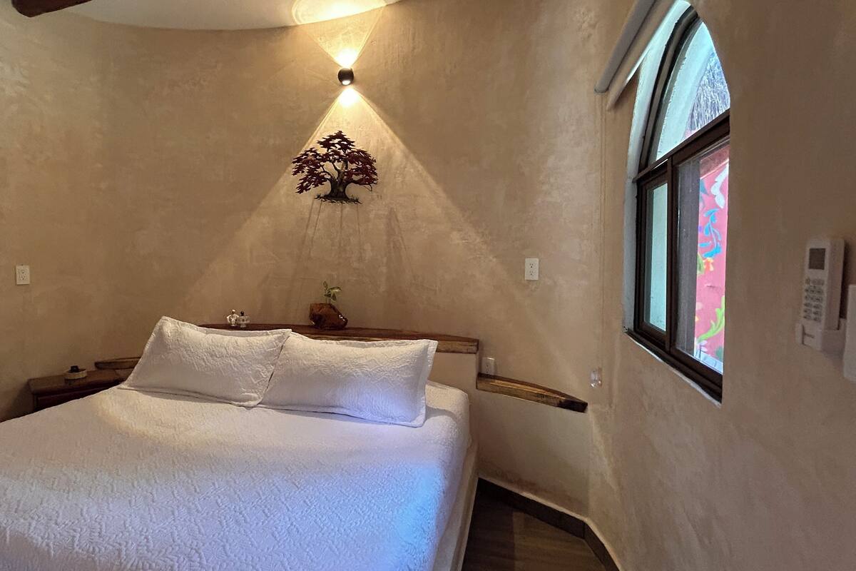 Un dormitorio con cama, mesita de noche, luz de pared, ventana y termostato en la pared.
