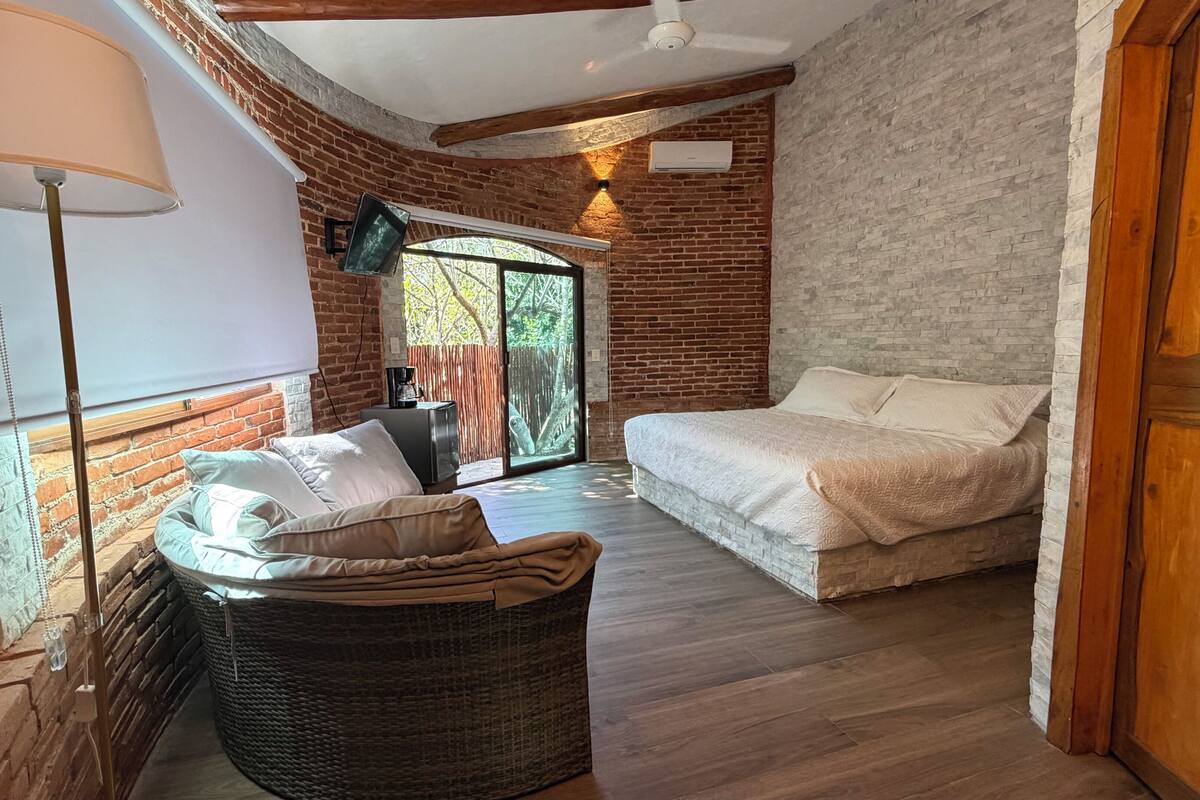 Una habitación acogedora con cama, una silla de mimbre, una lámpara y un ventanal con vistas a la vegetación.
