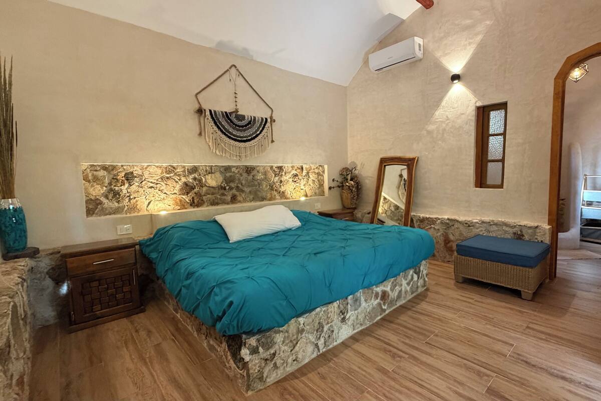 Un dormitorio con pared de piedra, una cama con colcha color esmeralda, una mesita de noche de madera, un banco azul y una puerta de madera que da a otra habitación.