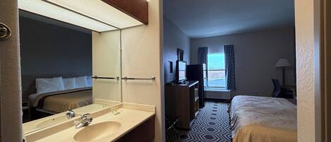 Ein Hotelzimmer mit großem Spiegel, Waschbecken, einem Bett mit gemustertem Überwurf, einem Fernseher und einem Fenster mit Vorhängen.