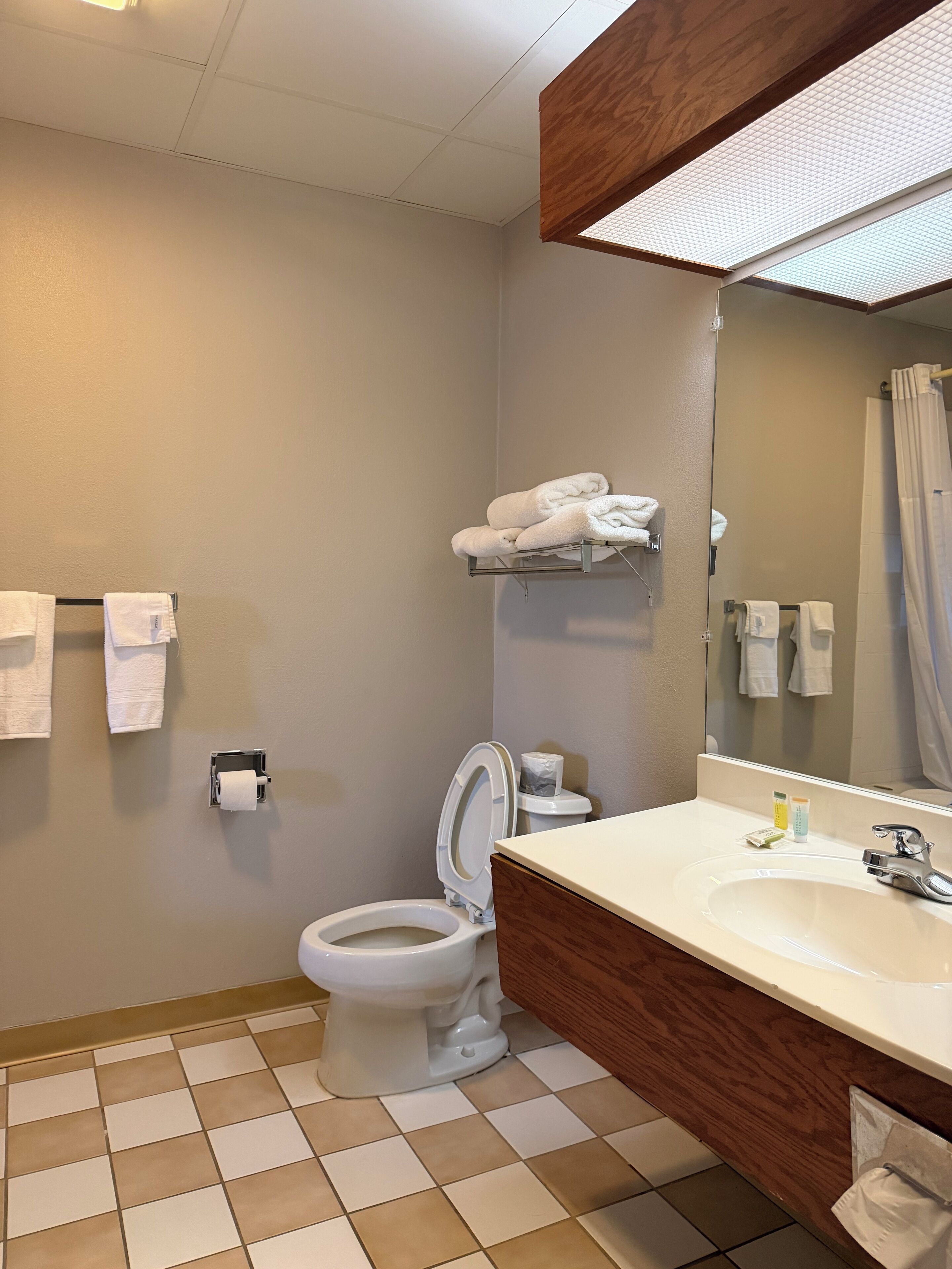 Un baño con inodoro, lavamanos y espejo. Hay toallas colgadas en la pared y una repisa con más toallas.