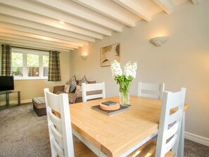 A dining area with a wooden table, white chairs, a vase with flowers, a sofa with plaid cushions, and a television on a stand. - 1 Clarkes Lane (Oswestry)