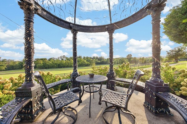 An outdoor gazebo with a table and chairs, surrounded by greenery and a view of the countryside.