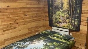 A wooden cabin bedroom with a bed featuring a forest-themed bedspread, a window with a forest view, and a hanging light fixture. - Chalet-style self-contained cottage in quiet countryside (Voiron)