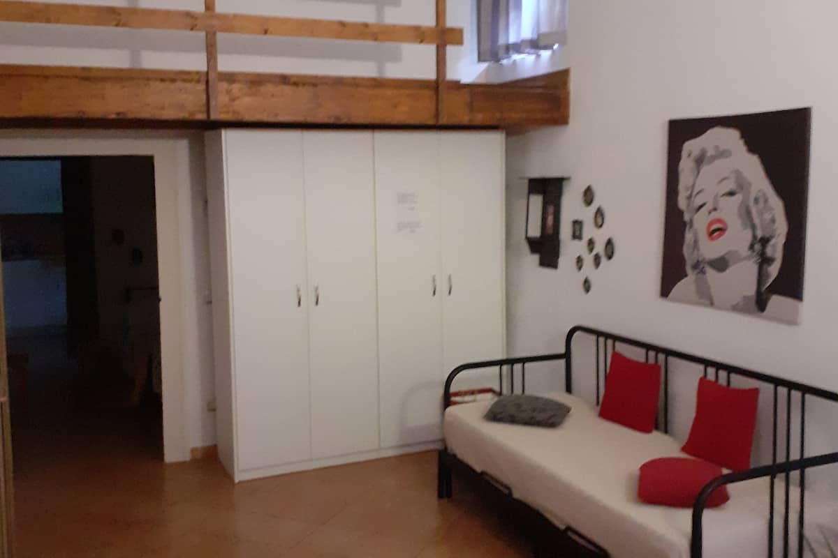 Um quarto com uma cama de solteiro, um sofá com almofadas vermelhas, uma parede com um quadro e um gato no chão.