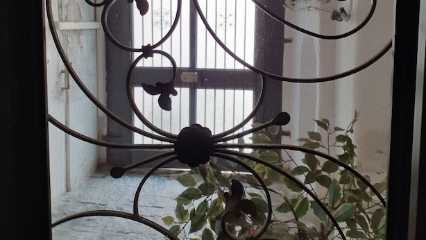 A wrought iron gate with floral designs, a window with vertical blinds, and a potted plant in the foreground. - casa normanna (Salerno)