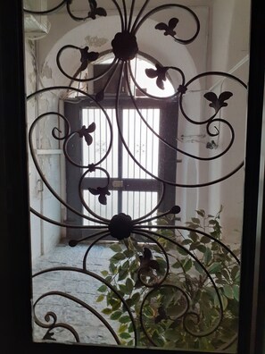 A wrought iron gate with floral designs, a window with vertical blinds, and a potted plant in the foreground.