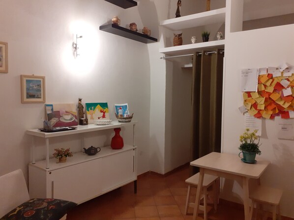 Una habitación pequeña y luminosa, con un escritorio blanco, una silla y una estantería con varios objetos decorativos. Se ve una puerta con cortinas y una pared con un tablón de anuncios.
