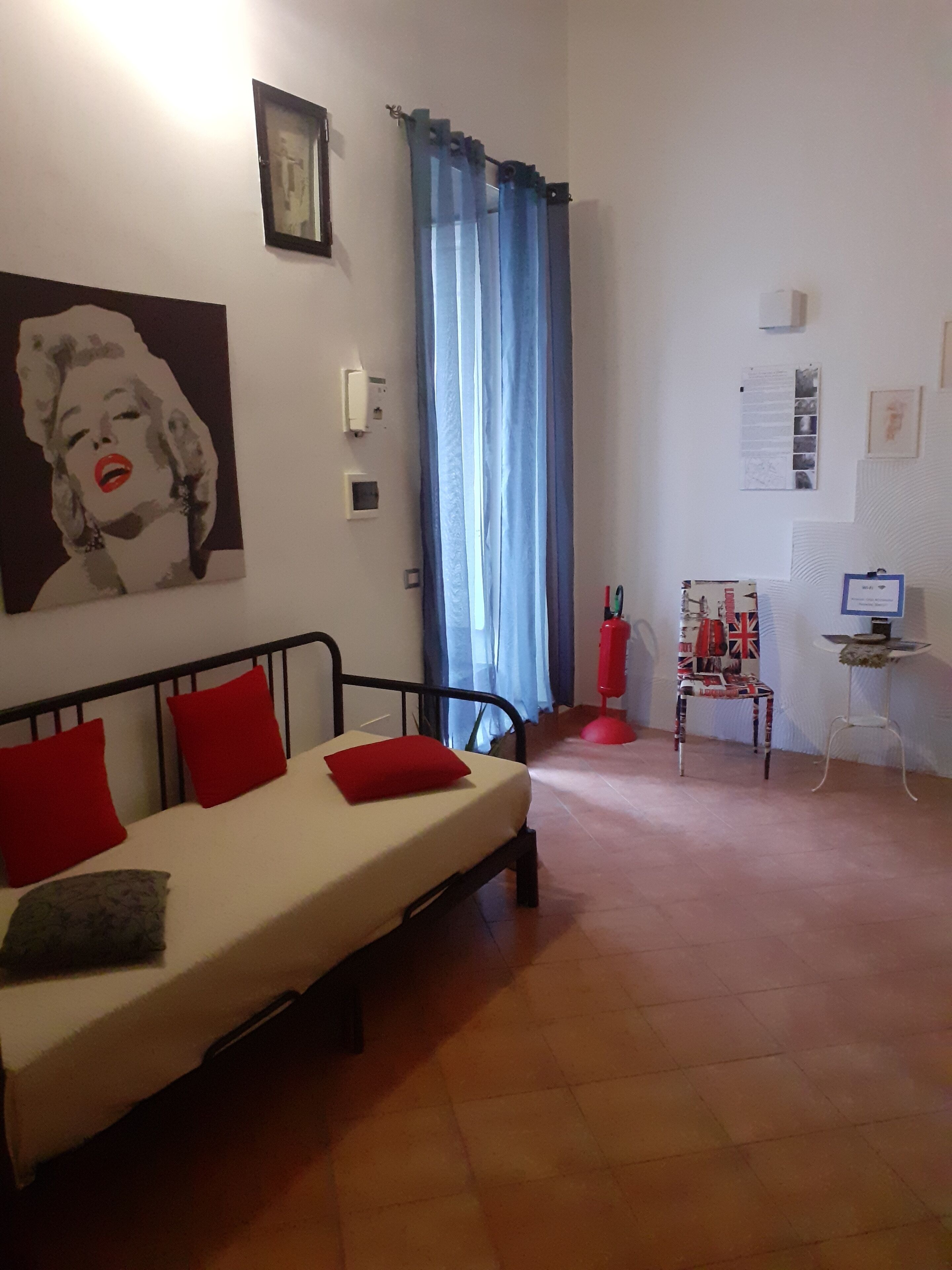 A bedroom with a bed, red pillows, a wall-mounted picture, a window with blue curtains, and a small table with a flag-patterned chair.