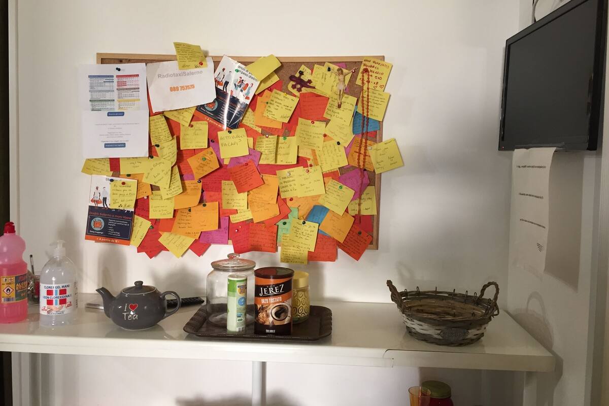 Um mural cheio de avisos e papéis fixados, uma prateleira com utensílios de cozinha, incluindo um bule, uma lata de café e um frasco de sabonete líquido.