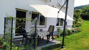 A patio with a table and chairs, a white umbrella, and a garden with flowers and plants.