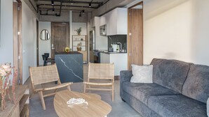 Ein modernes Wohnzimmer mit einem grauen Sofa, zwei Korbstühlen, einem Holztisch und einer Küche im Hintergrund.