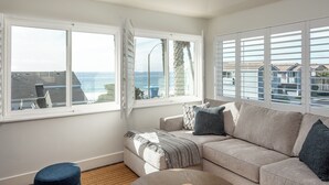 Ett vardagsrum med en beige soffa, en blå pall och utsikt över havet genom stora fönster.