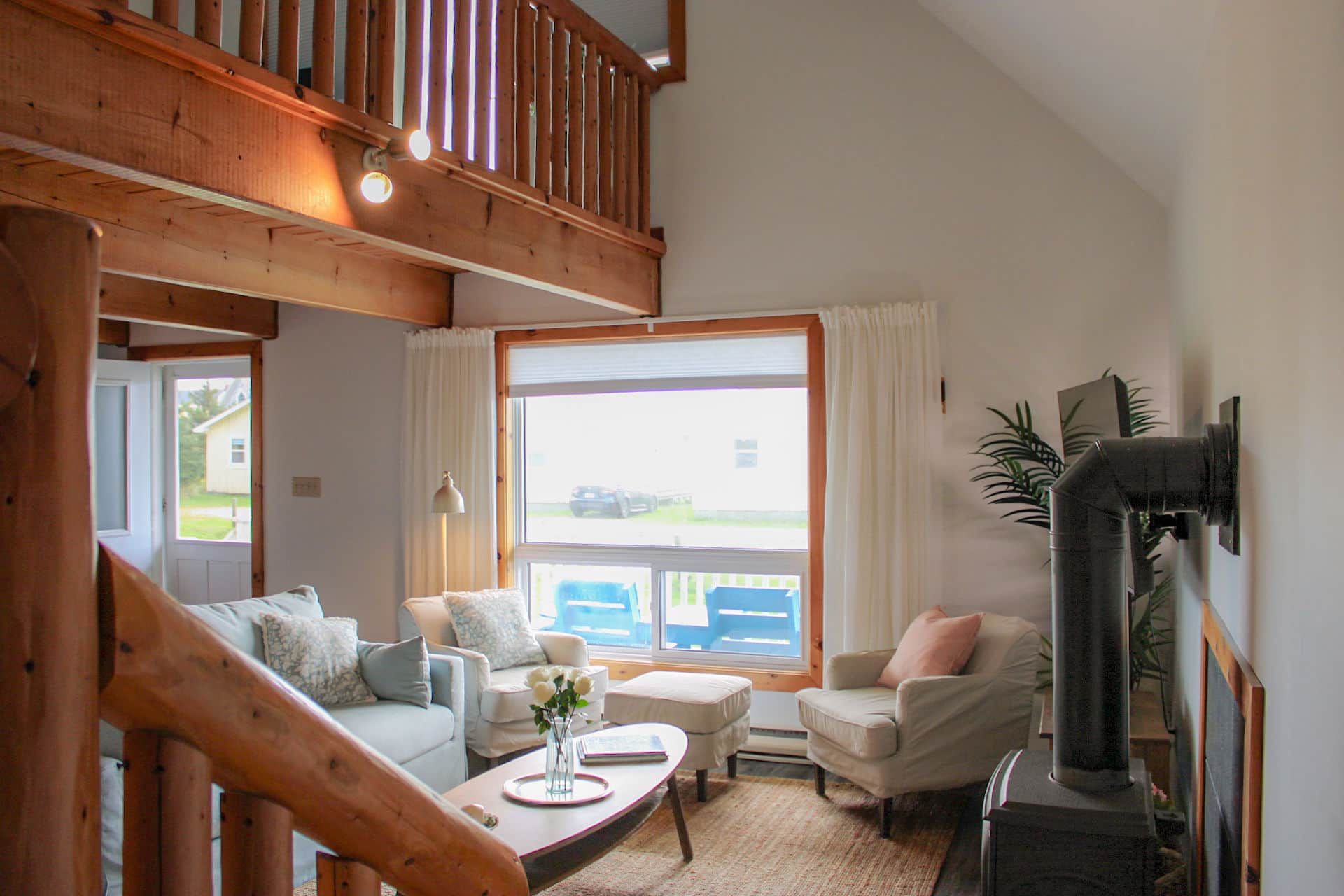 Un salon chaleureux avec un escalier en bois, une grande fenêtre avec des rideaux, une cheminée et des sièges confortables.