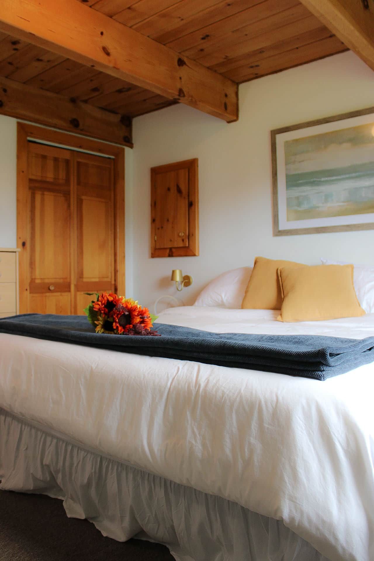 A hotel room with a bed, wooden ceiling beams, a wall-mounted light, and a framed picture.