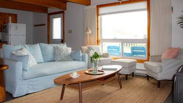 Ein Wohnzimmer mit einer Couch, einem Couchtisch und einem Sessel. Im Hintergrund ist ein Fenster mit Vorhängen, eine Lampe und ein Kühlschrank zu sehen.