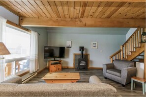 A cozy living room with a wooden ceiling, a television, a fireplace, and a staircase. - Ocean-View Chalet · King + 2 Queens · Pet-Friendly (Lockeport)