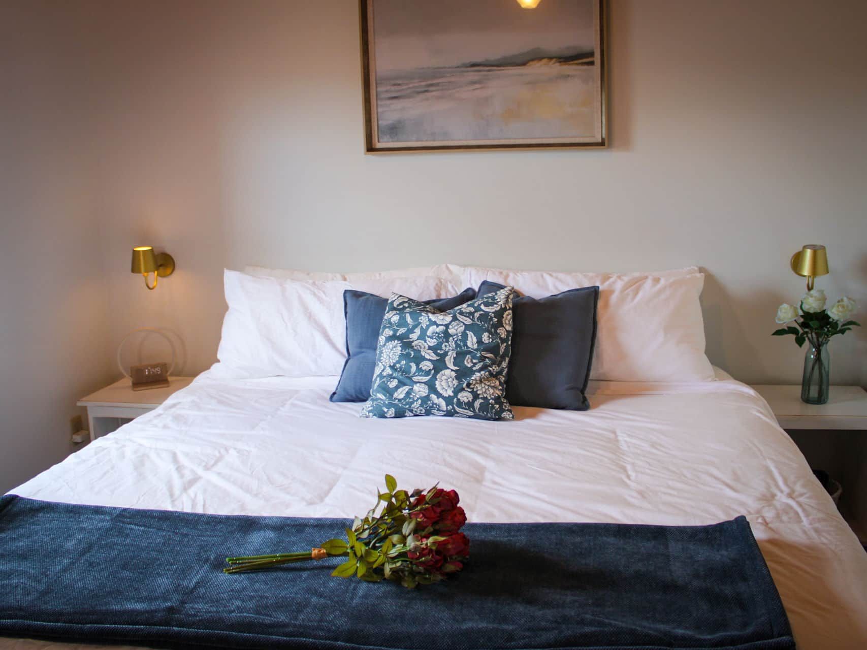 Una cama bien hecha con sábanas blancas, un cojín floral azul y un ramo de flores sobre una colcha azul.