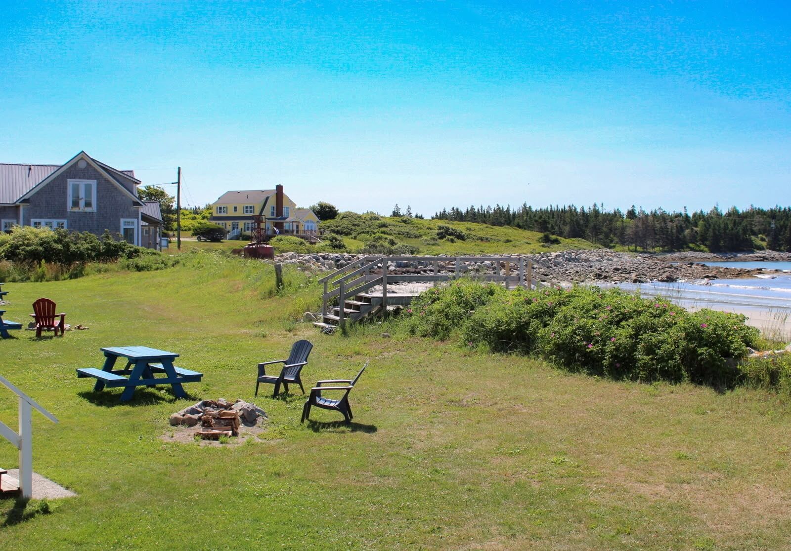A coastal property with a grassy area, picnic tables, and chairs, a stone staircase leading to a beach, and two houses in the background.