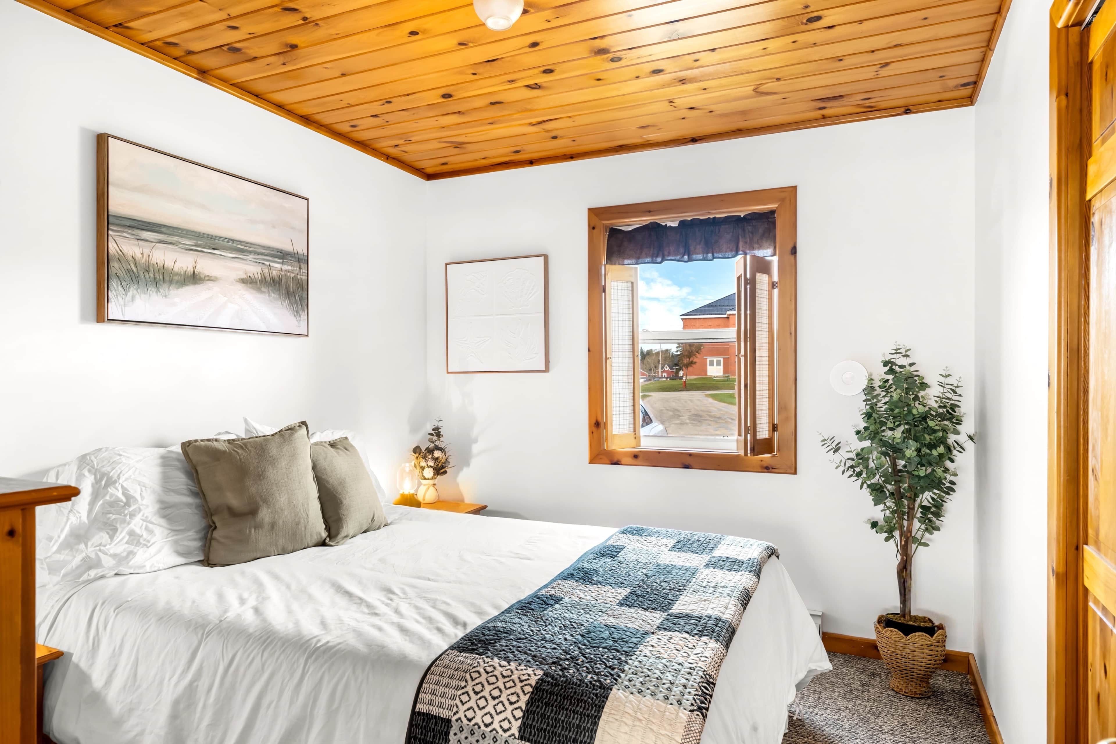 Een slaapkamer met een houten plafond, een bed met wit beddengoed, een sprei, een nachtkastje met een bloevas, een raam met uitzicht en een kamerplant.