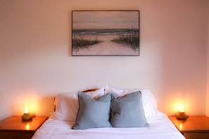 A bedroom with a bed, pillows, a painting of a beach, and two bedside lamps.