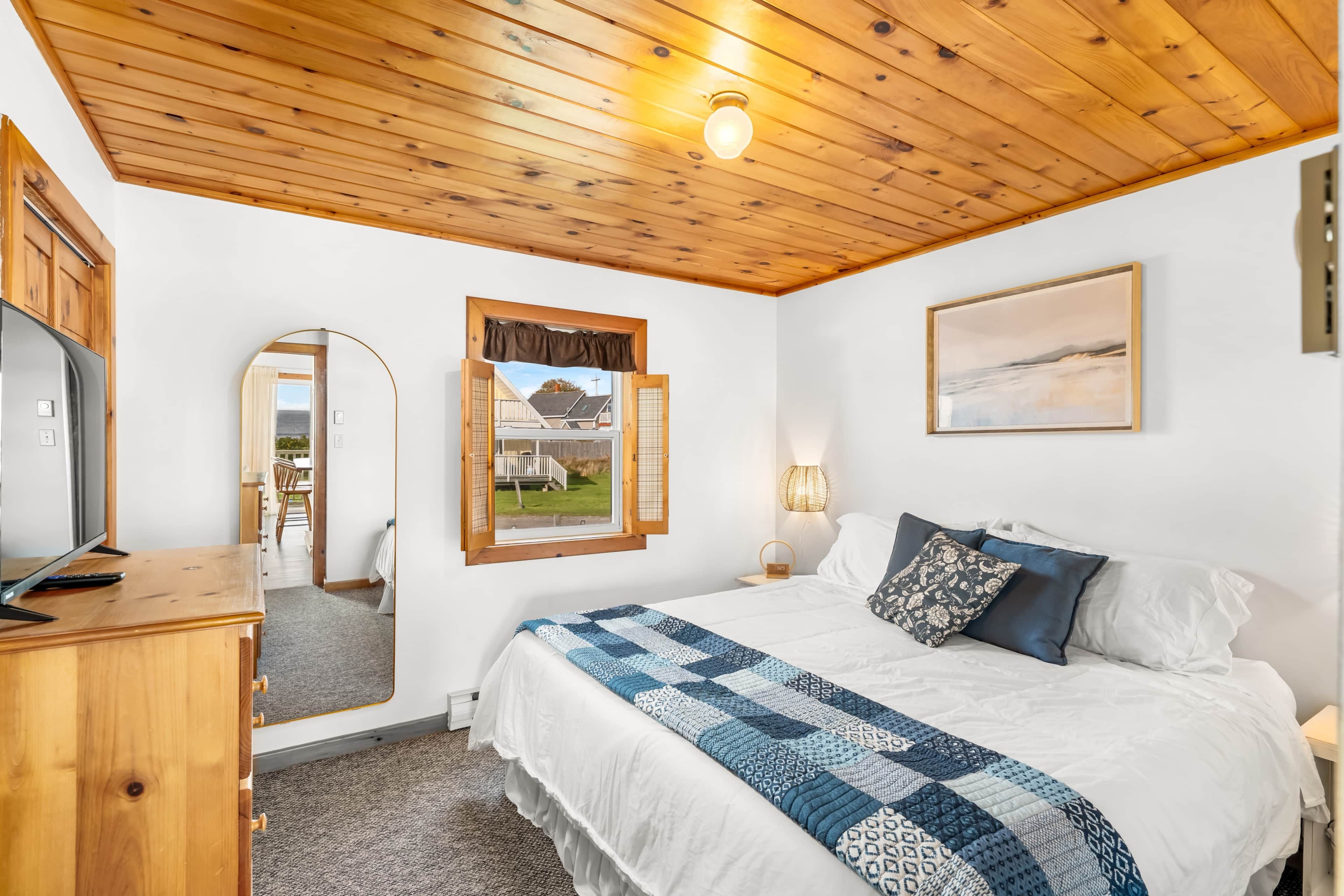 A bedroom with a wooden ceiling, a bed with a patterned quilt, a bedside lamp, a framed picture on the wall, and a view through a window.