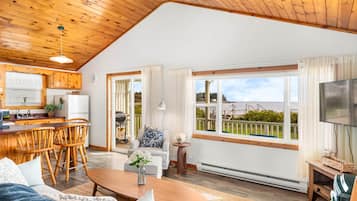 Un vaste espace de vie doté d’un plafond en bois, une cuisine avec des meubles en bois, un coin repas avec des chaises, un canapé, une table basse et une grande fenêtre offrant une vue sur l’extérieur.
