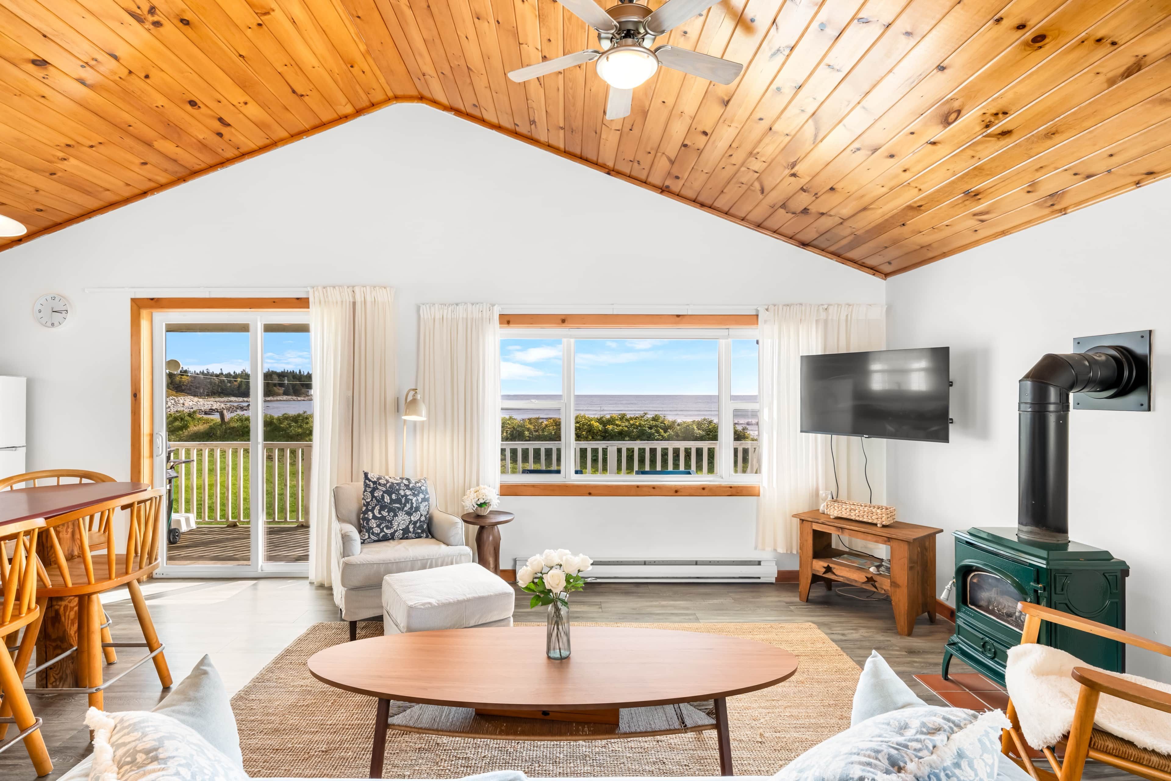 A spacious living room with a wooden ceiling, a large window with a view, a television, and a fireplace.