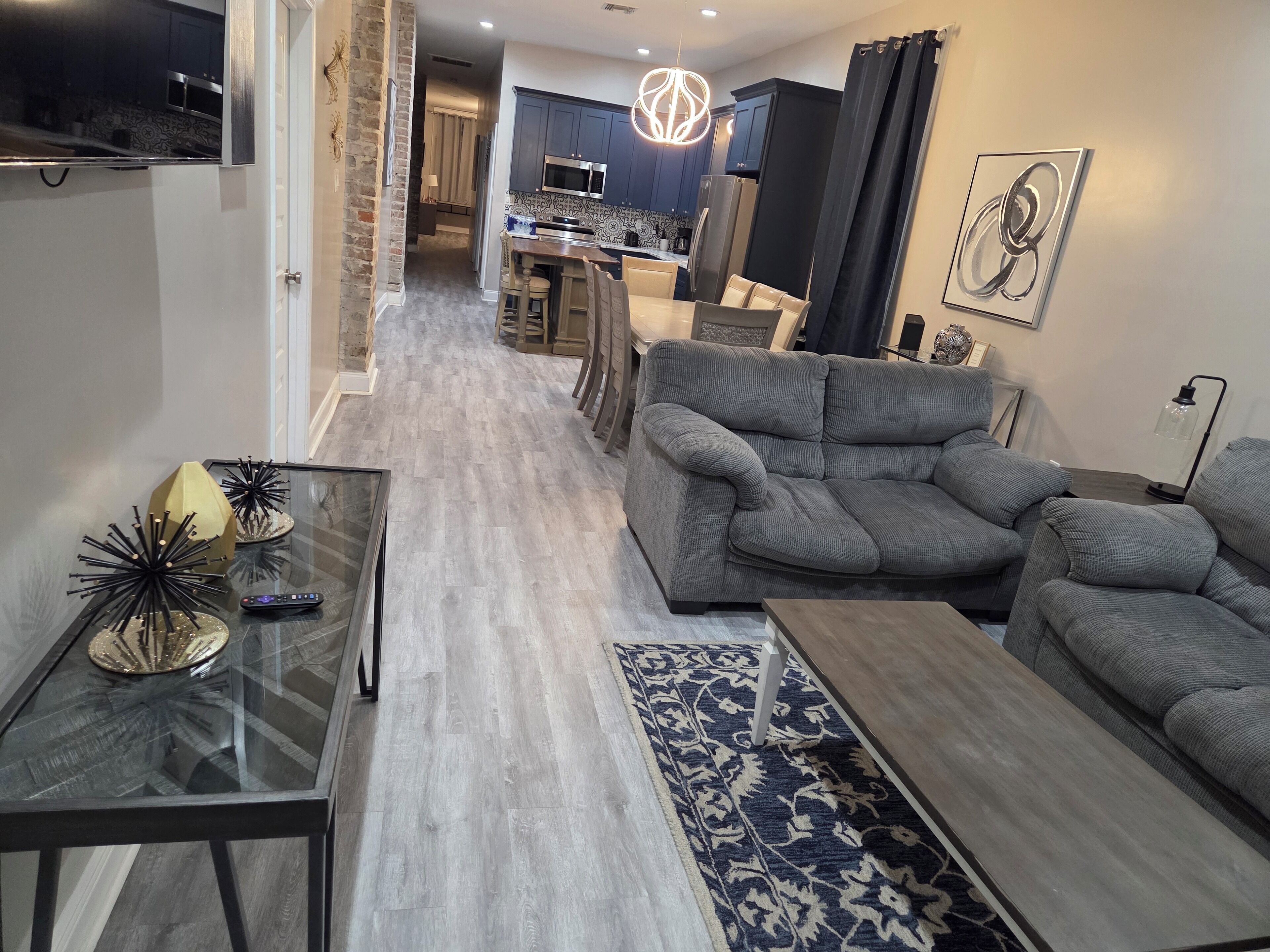 A modern living room with a grey sofa, a glass coffee table, and a patterned rug. A kitchen with dark cabinets and a microwave is visible in the background.